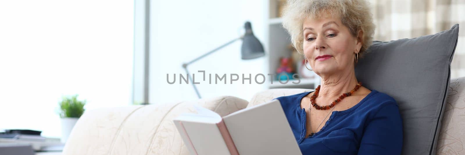 Elderly woman is reading book at home on couch. Home leisure pensioners concept