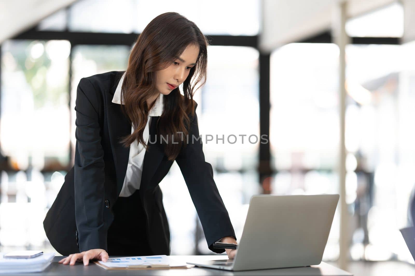 Focused young businesswoman standing at table in office, using laptop, looking at computer screen, reading or writing business email, searching information in internet, working on project..