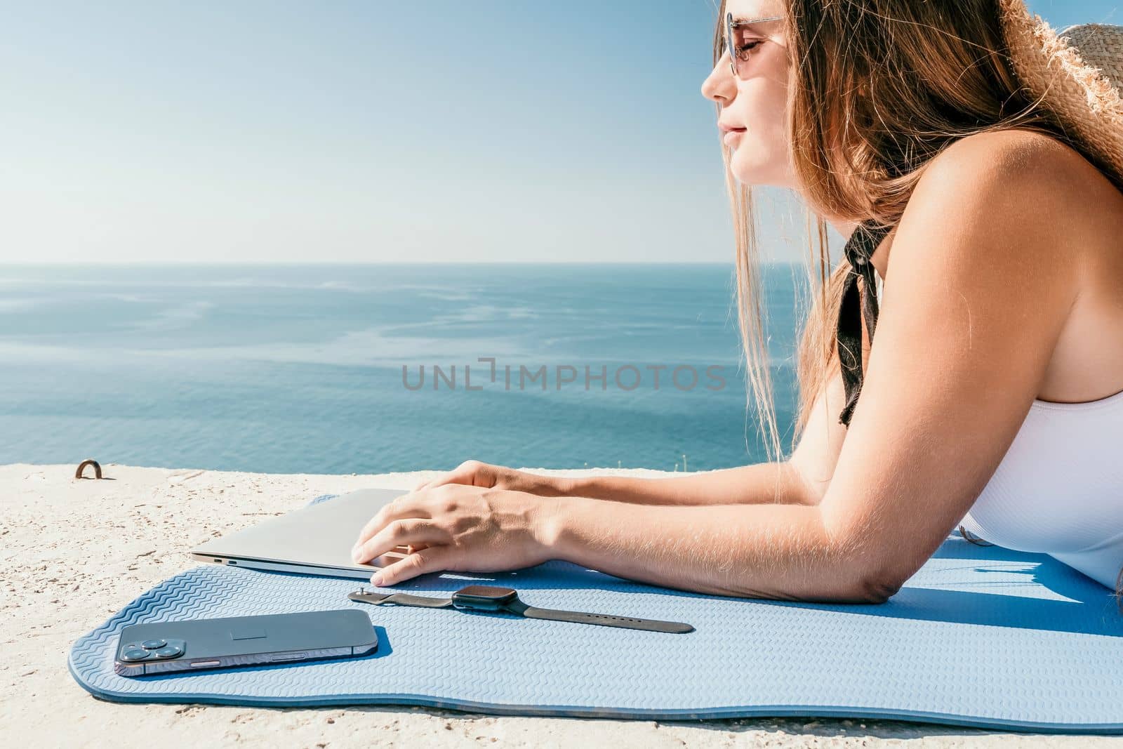 Successful business woman in yellow hat working on laptop by the sea. Pretty lady typing on computer at summer day outdoors. Freelance, travel and holidays concept.