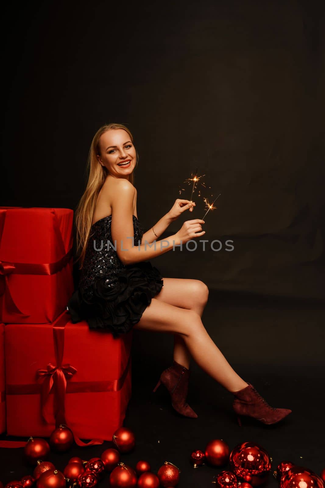 Woman gifts christmas. Beautiful sexy young blonde in a black dress sits on three big red gifts on a black background. Christmas photo, gifts.