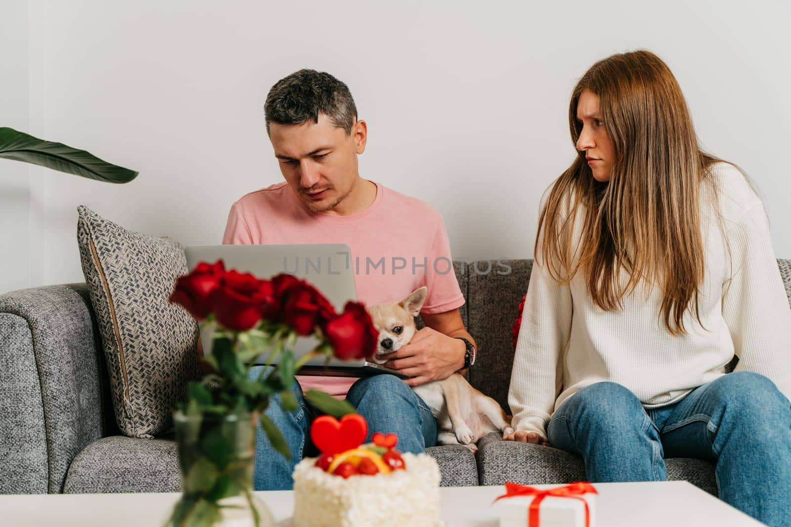 Beautiful young couple with dog sitting on a couch, having a quarrel over romantic date on Valentine day. Man working at laptop, woman in anger by Ostanina