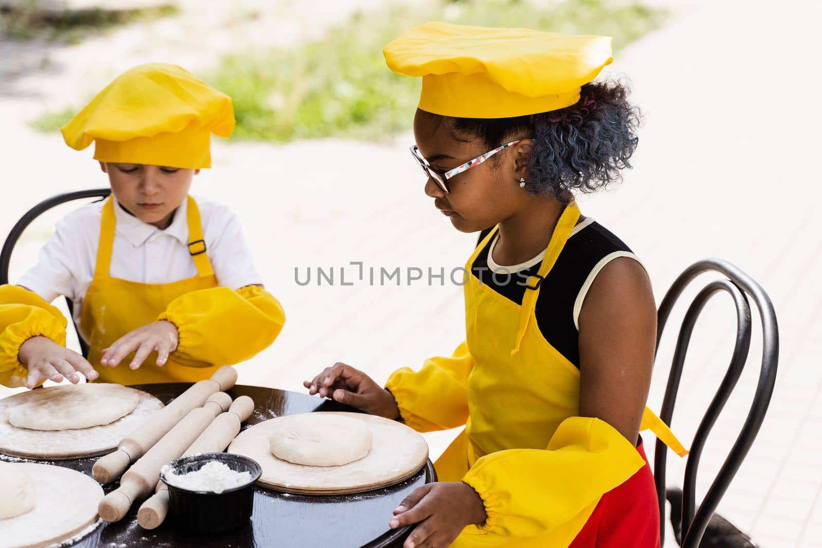 Multinational company of children cooks in yellow uniforms cooking dough for bakery. African teenager and black girl have fun with caucasian child boy and cook food. by Rabizo