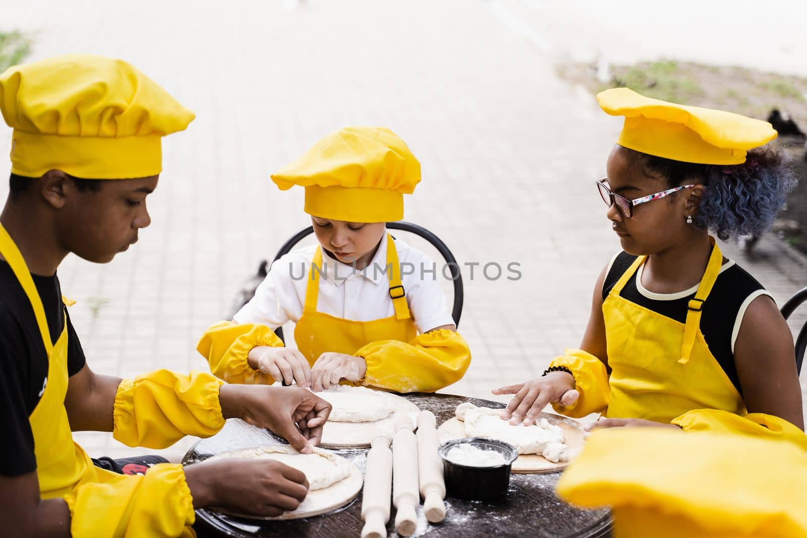 Multiethnic cooks children in yellow chefs hat and apron cooking dough for bakery. Black african and caucasian child cooking and having fun together