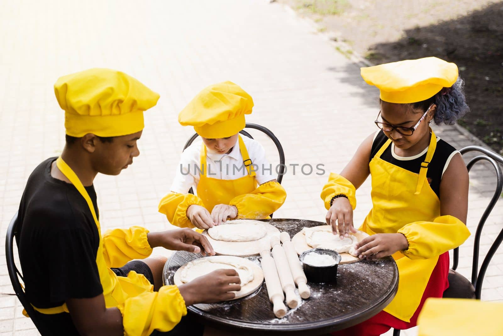 Multinational company of children cooks in yellow uniforms cooking dough for bakery. African teenager and black girl have fun with caucasian child boy and cook food