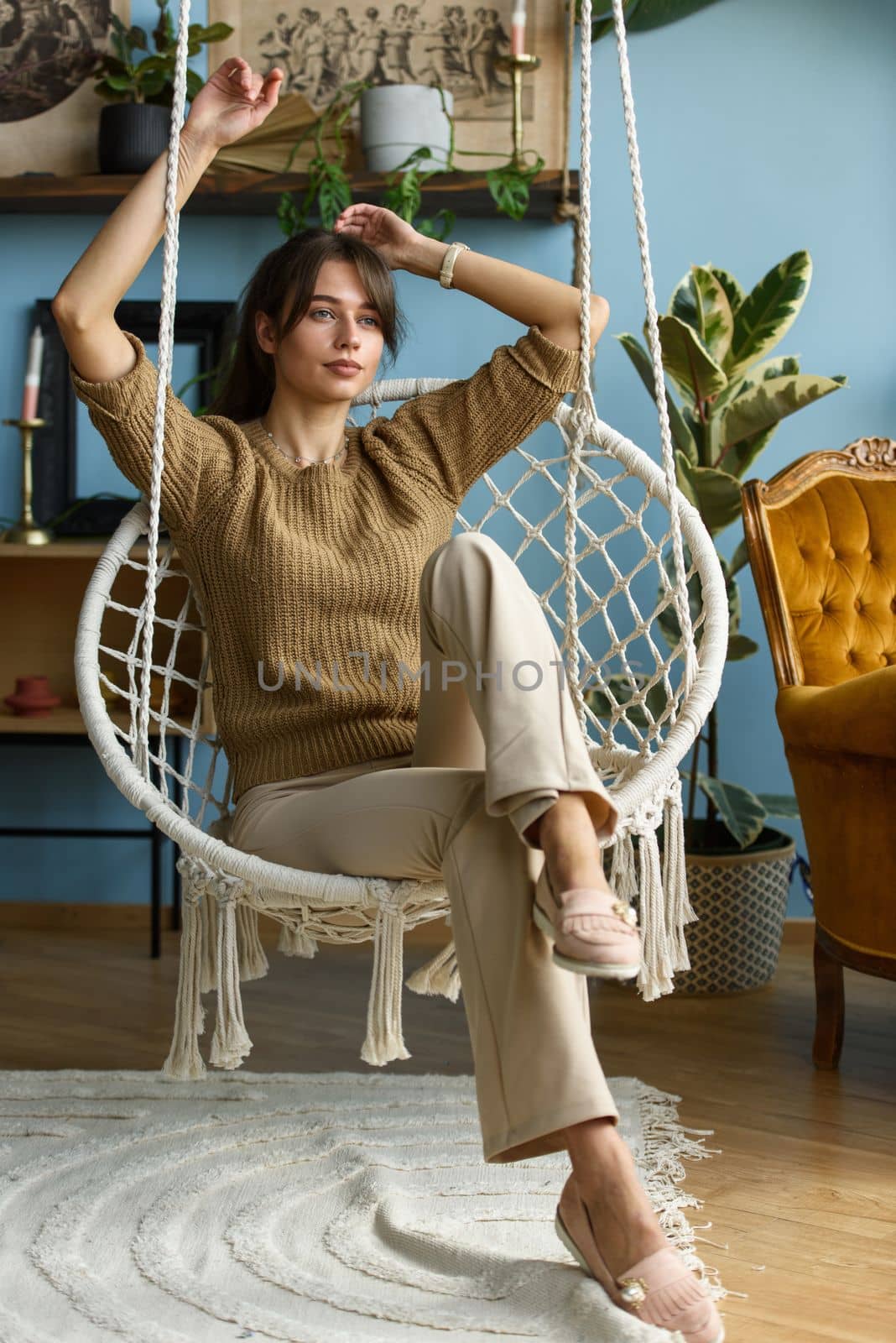 Young attractive woman chilling at home in comfortable hanging chair. beautiful slender girl in beige pants and olive sweater posing while sitting in a swing chair