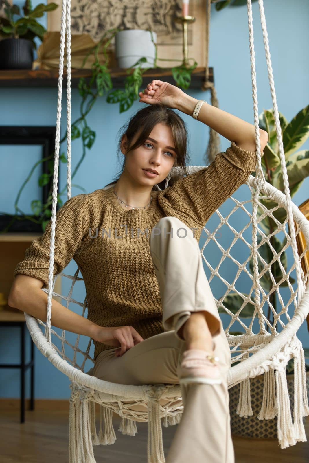 Young attractive woman chilling at home in comfortable hanging chair. beautiful slender girl in beige pants and olive sweater posing while sitting in a swing chair