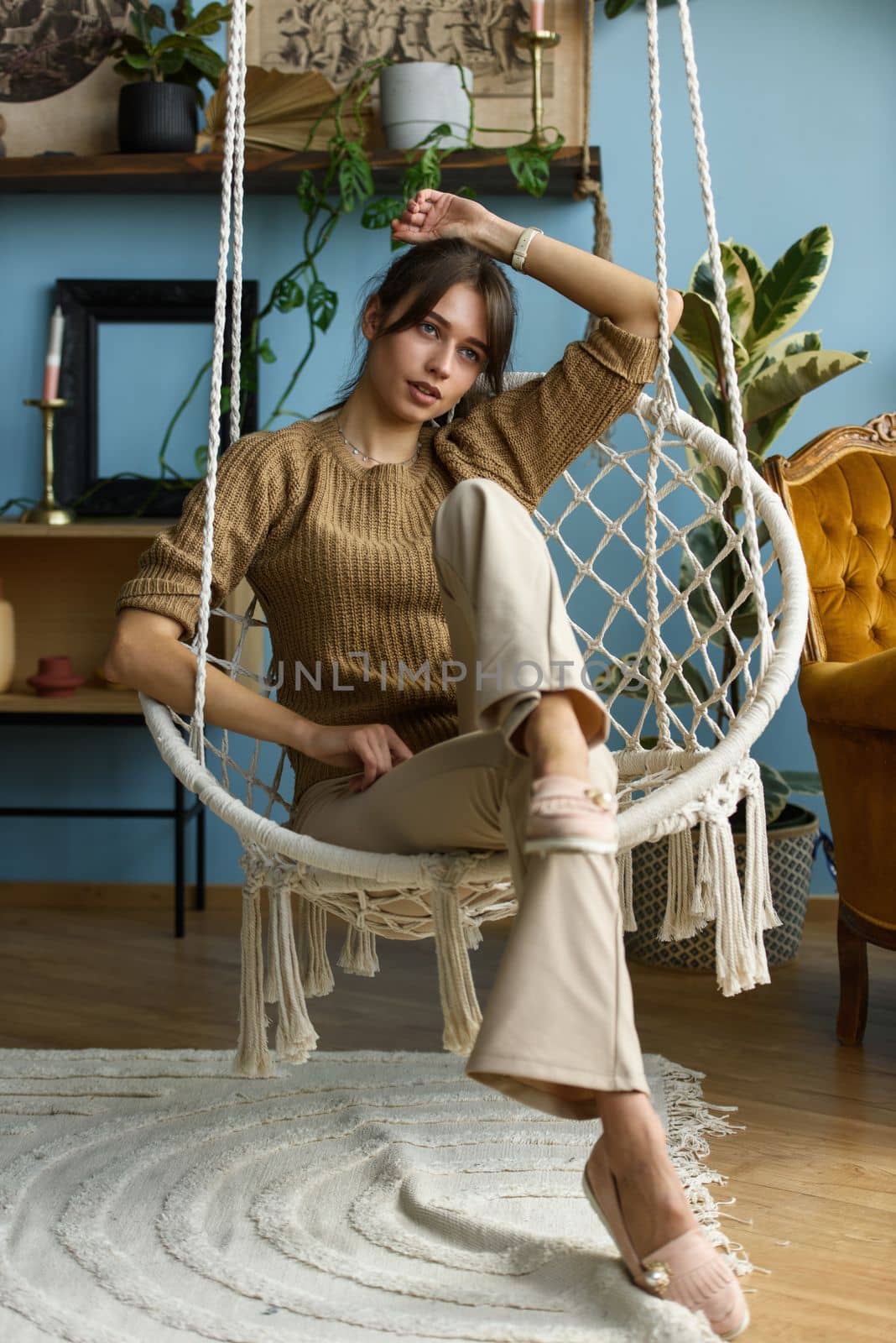 Young attractive woman chilling at home in comfortable hanging chair. beautiful slender girl in beige pants and olive sweater posing while sitting in a swing chair
