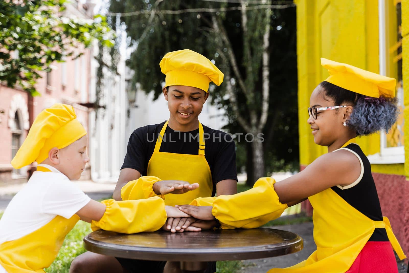 Teambuilding of multinational children cooks in chefs hat and yellow apron uniform put hands on each other, having fun and laughing. Multiethnic kids commutication activity. by Rabizo