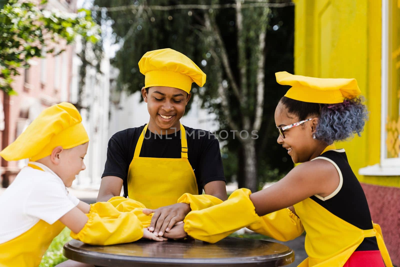 Teambuilding of multinational children cooks in chefs hat and yellow apron uniform put hands on each other, having fun and laughing. Multiethnic kids commutication activity