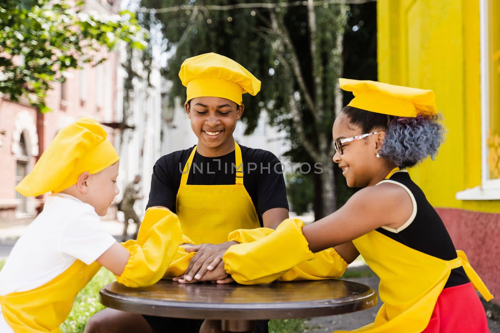 Teambuilding of multinational children cooks in chefs hat and yellow apron uniform put hands on each other, having fun and laughing. Multiethnic kids commutication activity