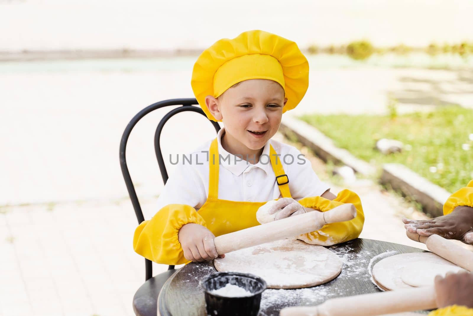 Handsome cook child in yellow chefs hat and apron yellow uniform holding dough roller and cooking dough outdoor