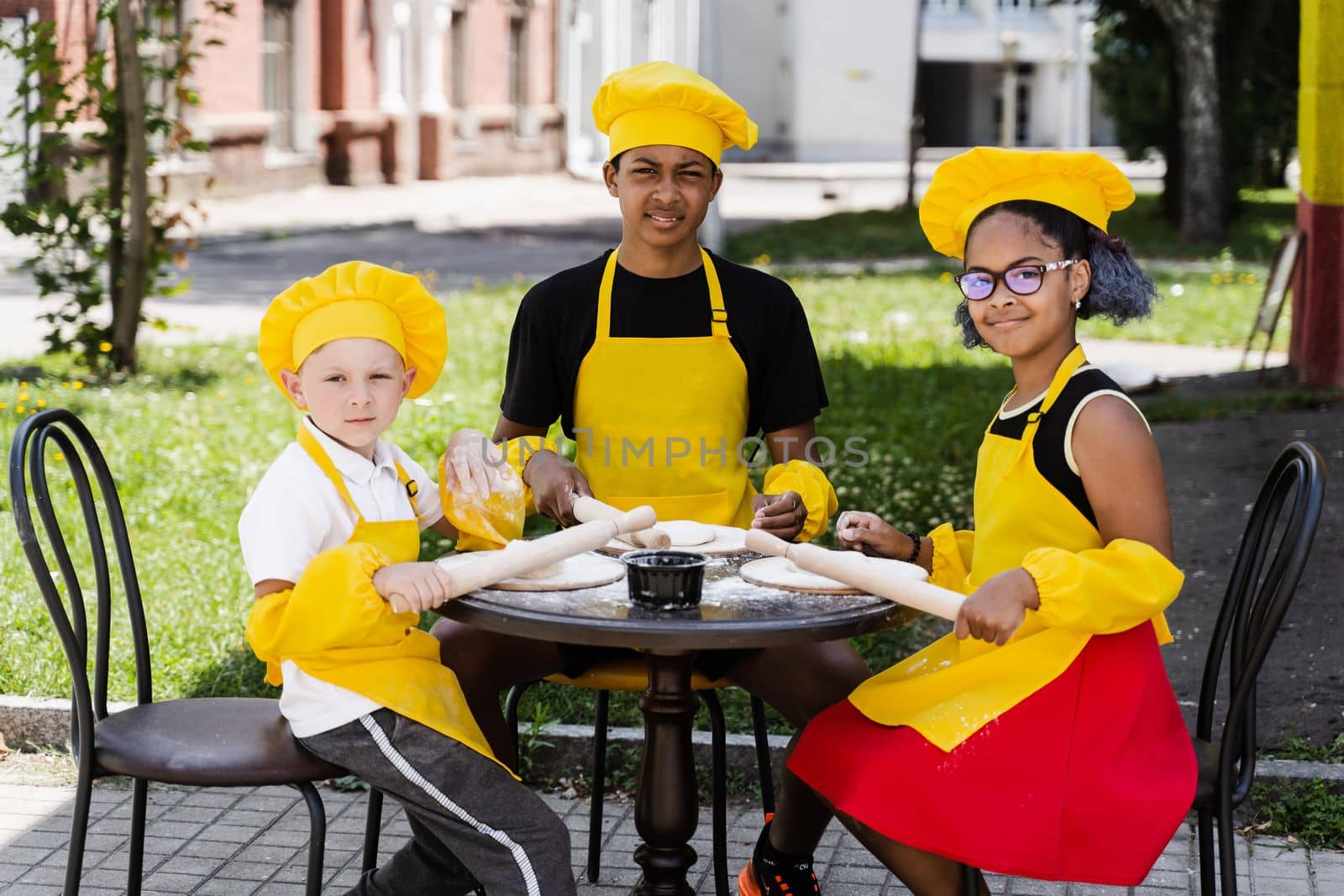 Multinational company of children cooks in yellow uniforms cooking dough for bakery. African teenager and black girl have fun with caucasian child boy and cook food. by Rabizo