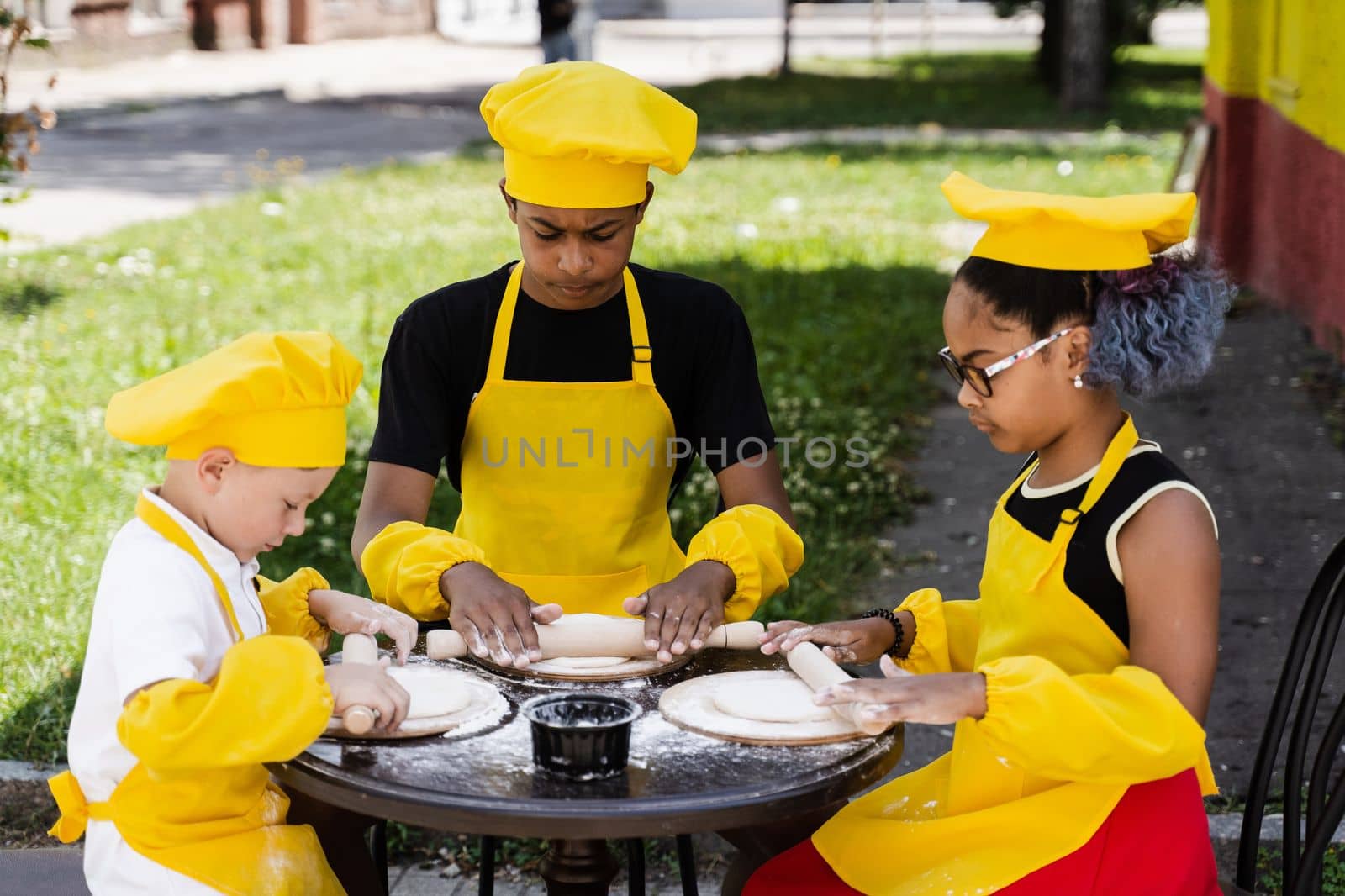 Multinational company of children cooks in yellow uniforms cooking dough for bakery. African teenager and black girl have fun with caucasian child boy and cook food