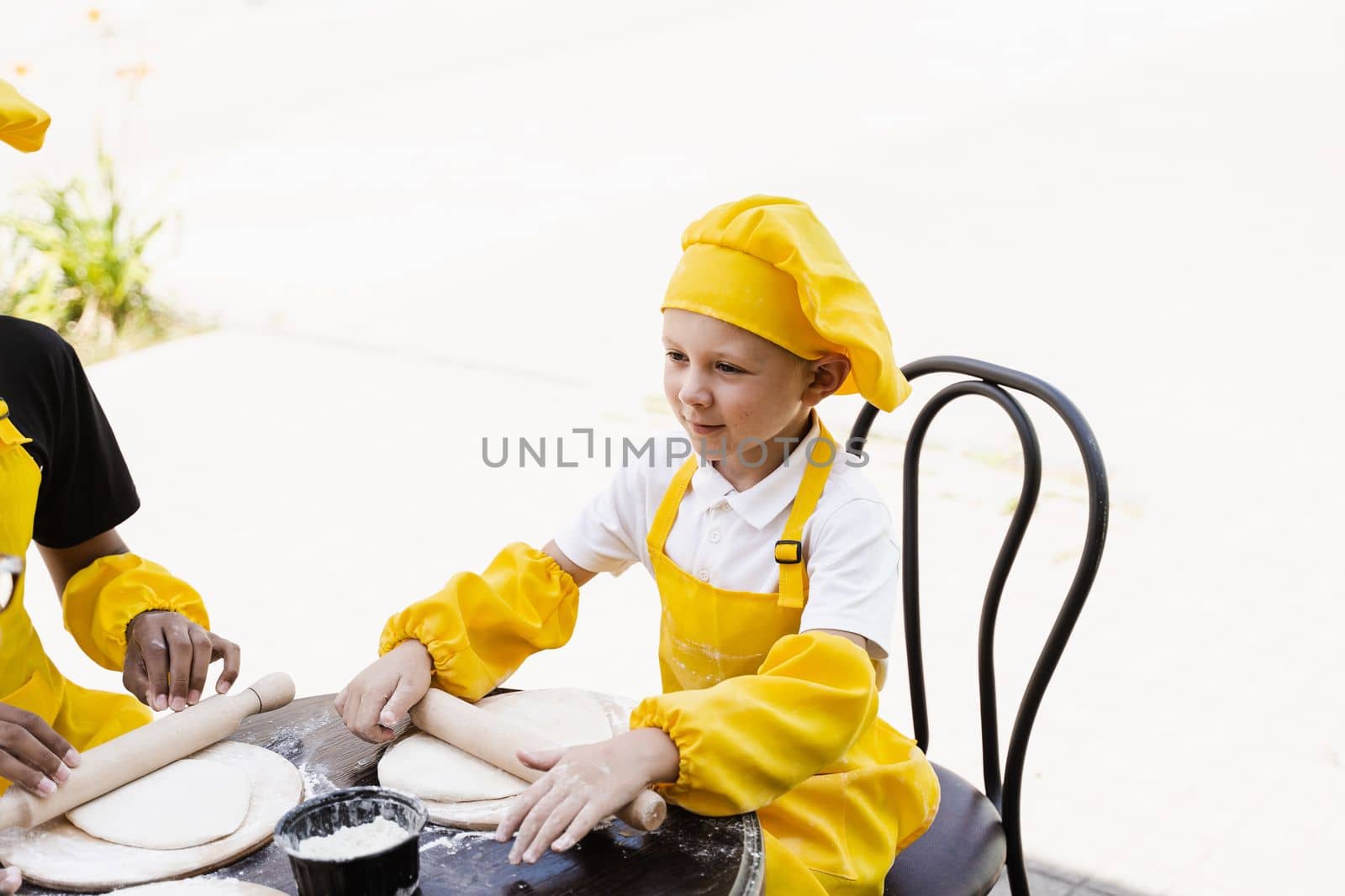 Handsome cook child in yellow chefs hat and apron yellow uniform rolling dough and cooking outdoor. by Rabizo