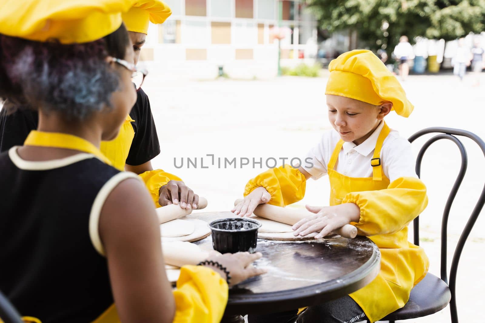 Handsome cook child in yellow chefs hat and apron yellow uniform rolling dough and cooking outdoor. by Rabizo