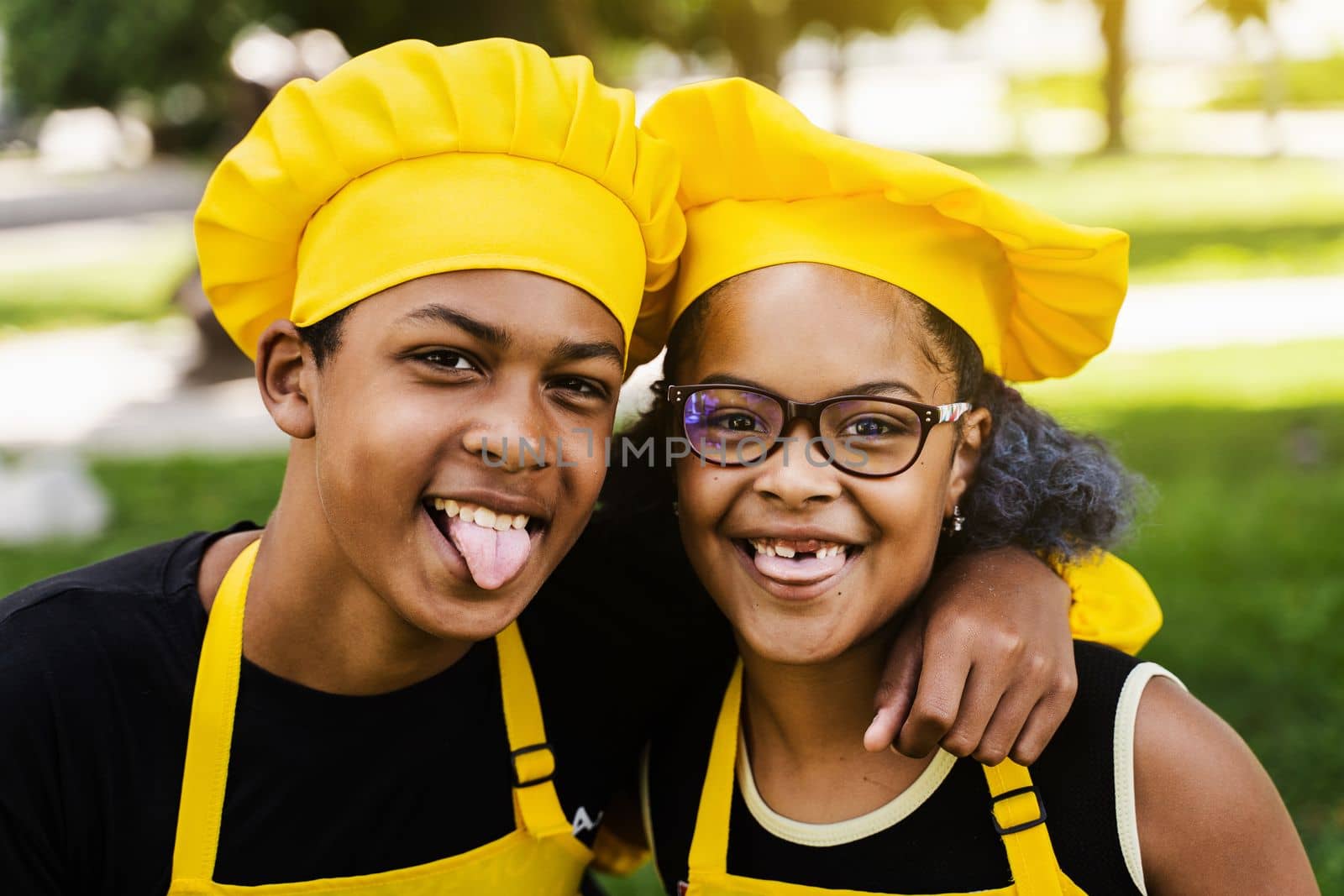African children cooks in chefs hat and yellow uniforms grimacing and showing tongue each others. African teenager and black girl have fun and cook food. by Rabizo