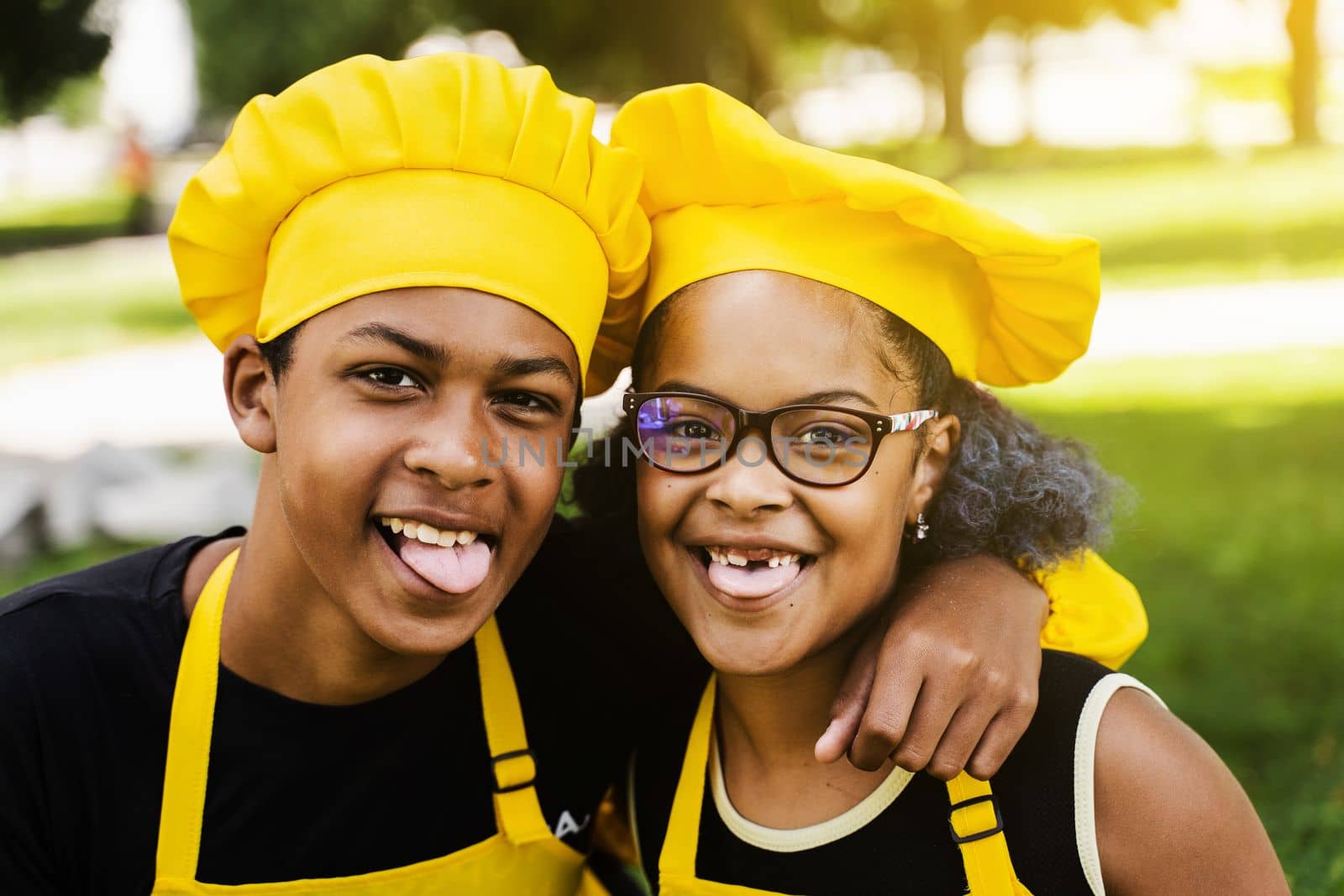 African children cooks in chefs hat and yellow uniforms grimacing and showing tongue each others. African teenager and black girl have fun and cook food