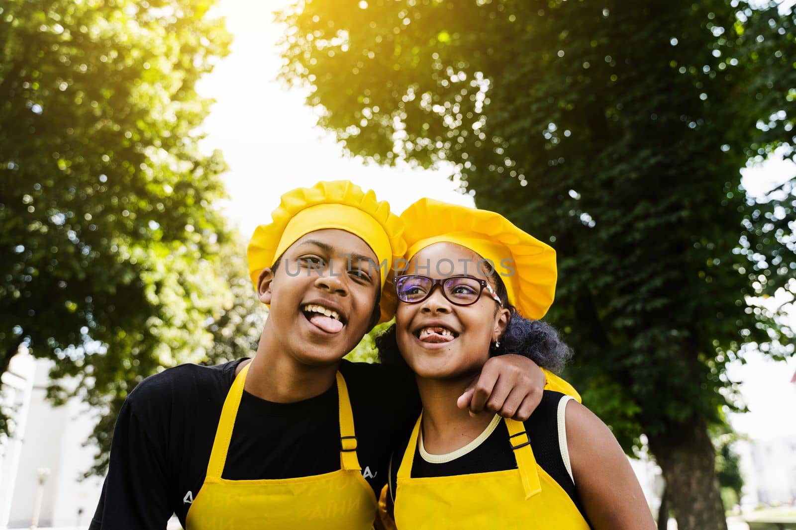 African children cooks in chefs hat and yellow uniforms grimacing and showing tongue each others. African teenager and black girl have fun and cook food. by Rabizo
