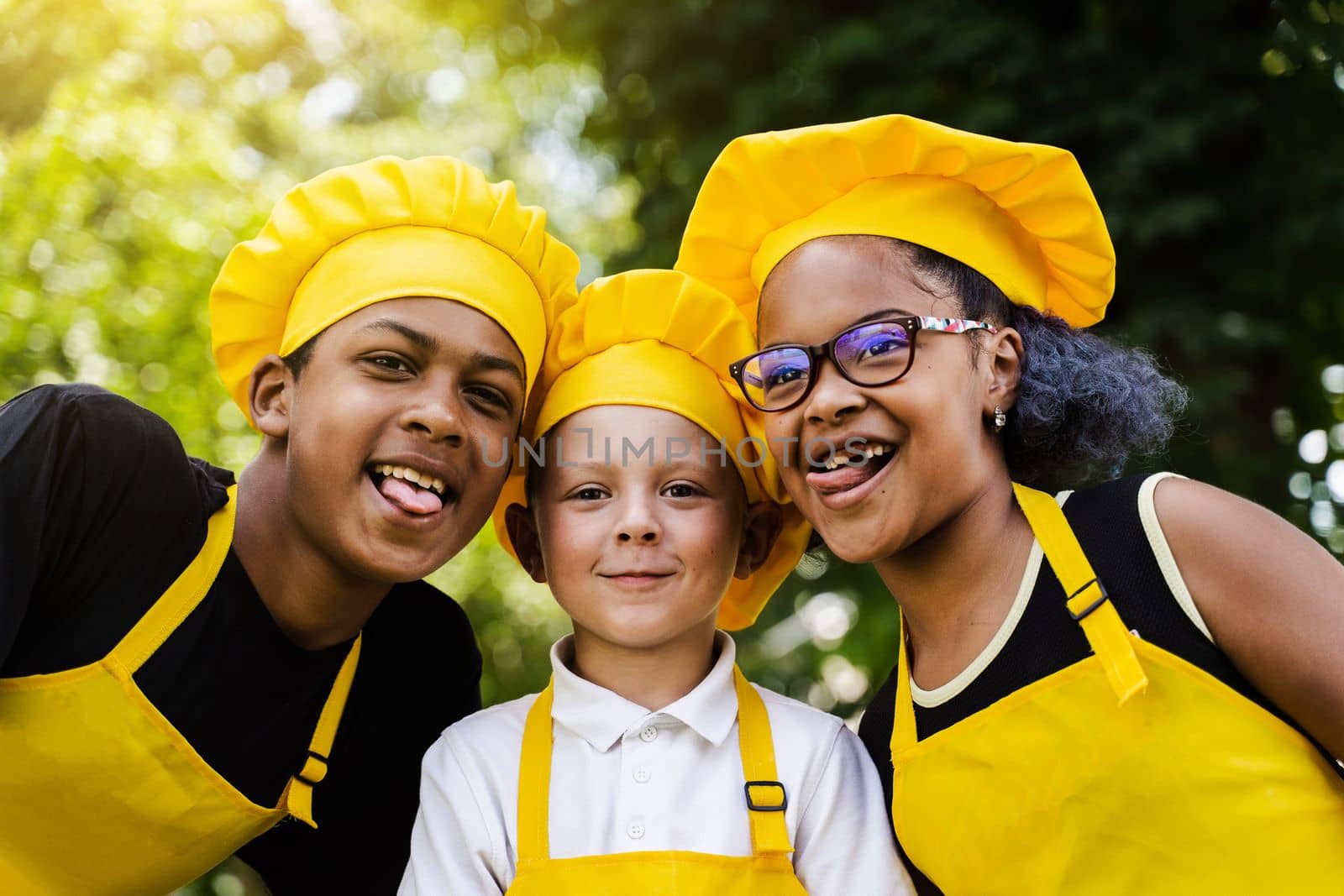 Multinational company of children cooks in yellow uniforms smiling and grimacing and showing tongue outdoor. African teenager and black girl have fun with caucasian child boy and cook food. by Rabizo