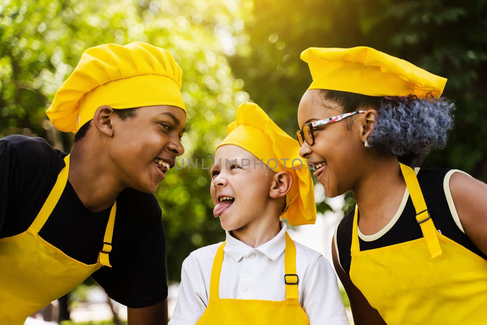 Multinational company of children cooks in yellow uniforms smiling and grimacing and showing tongue outdoor. African teenager and black girl have fun with caucasian child boy and cook food. by Rabizo