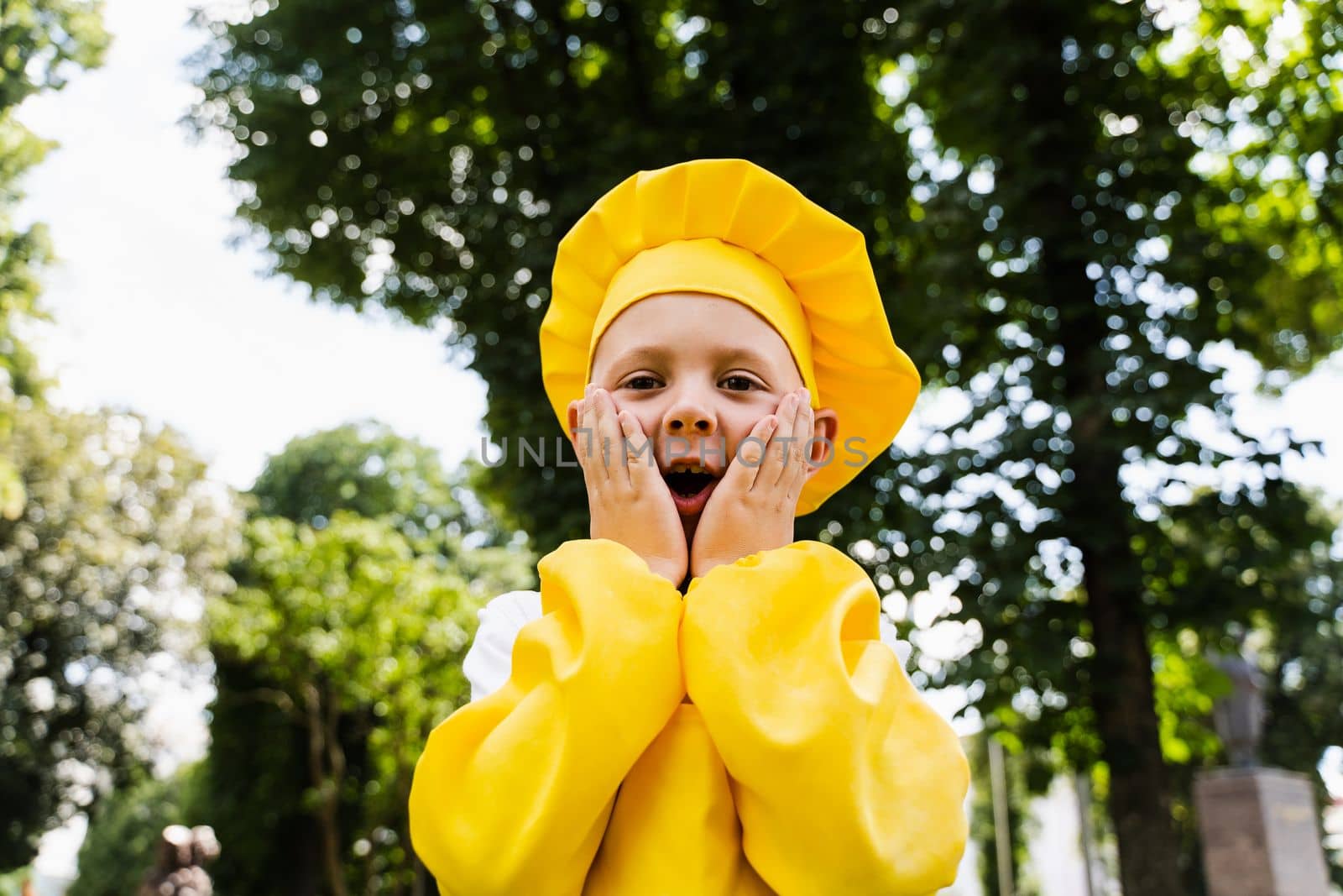 Shocked cook child in yellow chefs hat and apron yellow uniform holding his cheeks and surprise outdoor. by Rabizo
