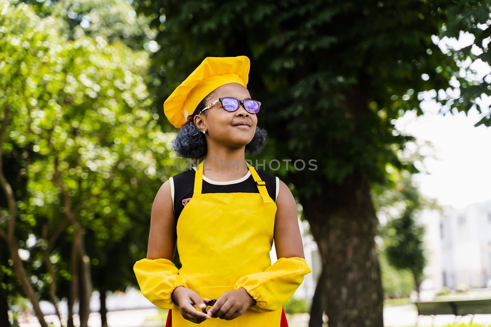 Black african child cook girl in chefs hat and yellow apron uniform smiling outdoor. Creative advertising for cafe or restaurant