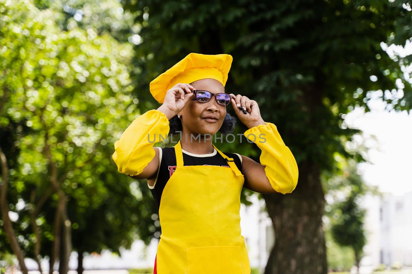 Black african child cook girl in chefs hat and yellow apron uniform smiling outdoor. Creative advertising for cafe or restaurant. by Rabizo