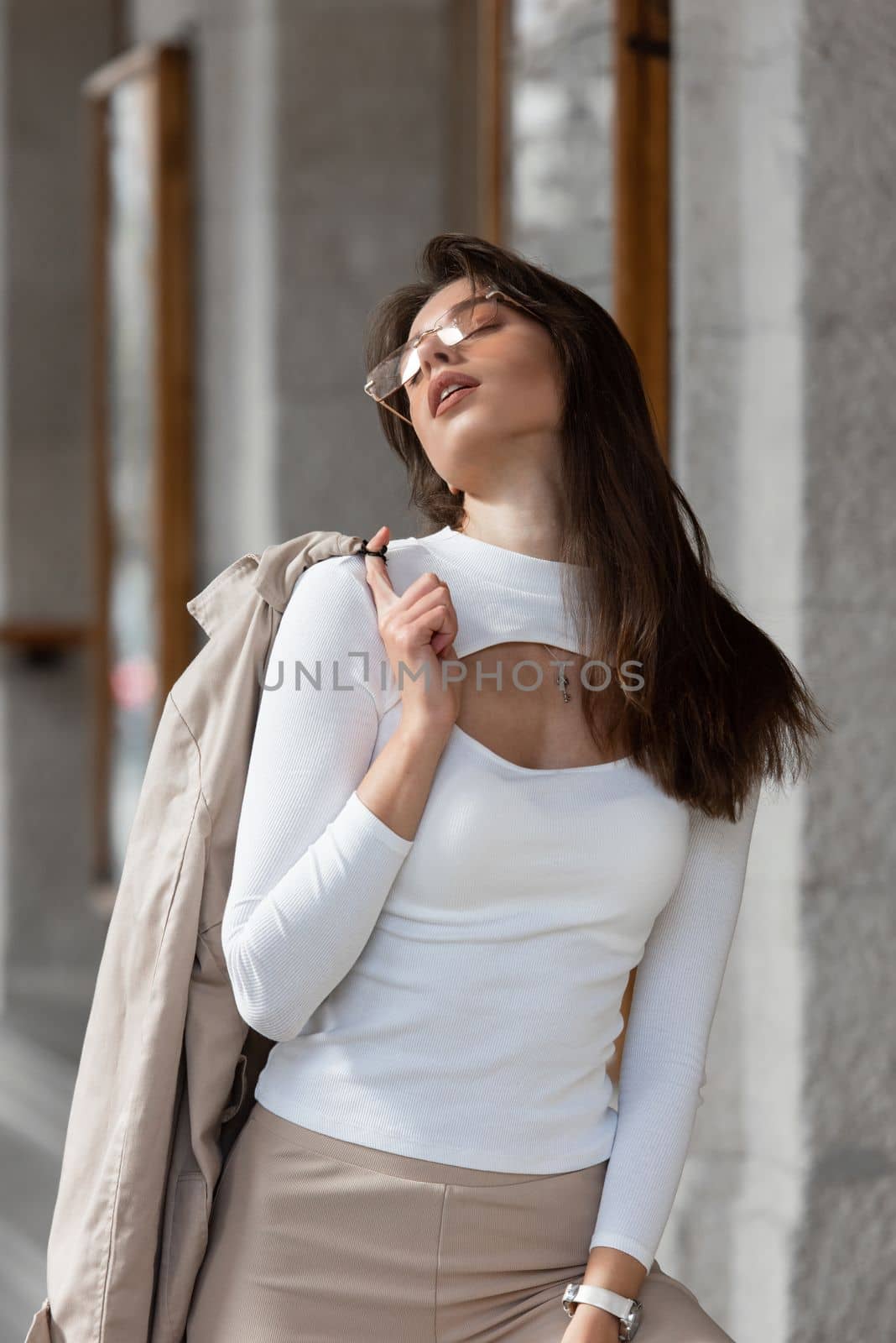 beautiful brunette girl in a sunglasses dressed in white blouse with a beige coat in hands. Stylish trendy fashion outlook by Ashtray25