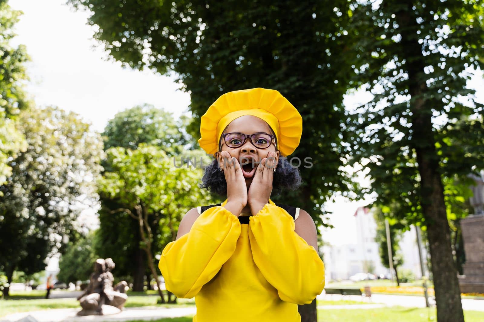 Shocked black african child cook girl in chefs hat and yellow apron uniform hold cheeks and surprise. Creative advertising for cafe or restaurant