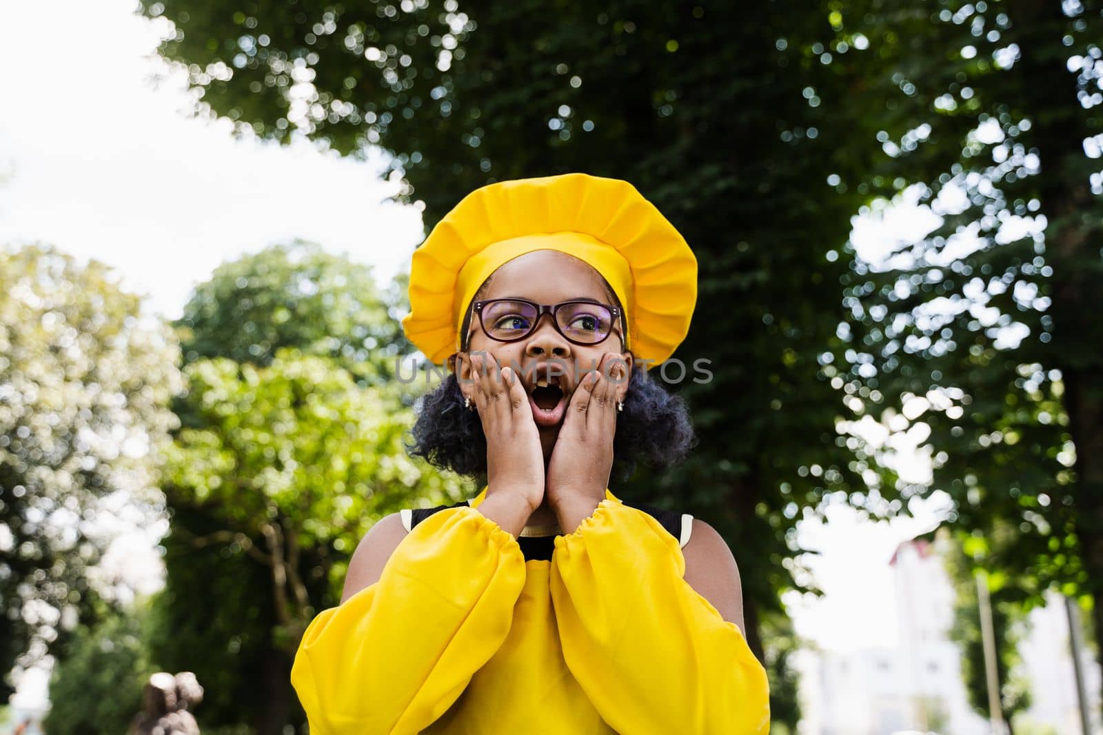 Shocked black african child cook girl in chefs hat and yellow apron uniform hold cheeks and surprise. Creative advertising for cafe or restaurant