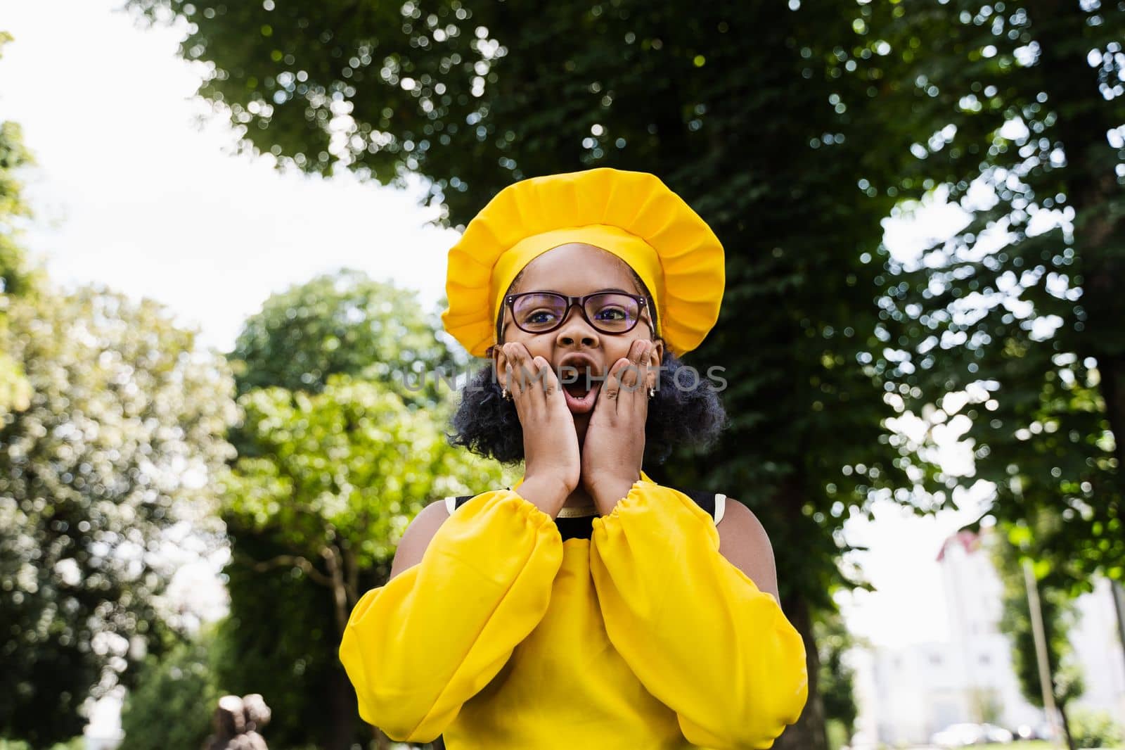 Shocked black african child cook girl in chefs hat and yellow apron uniform hold cheeks and surprise. Creative advertising for cafe or restaurant