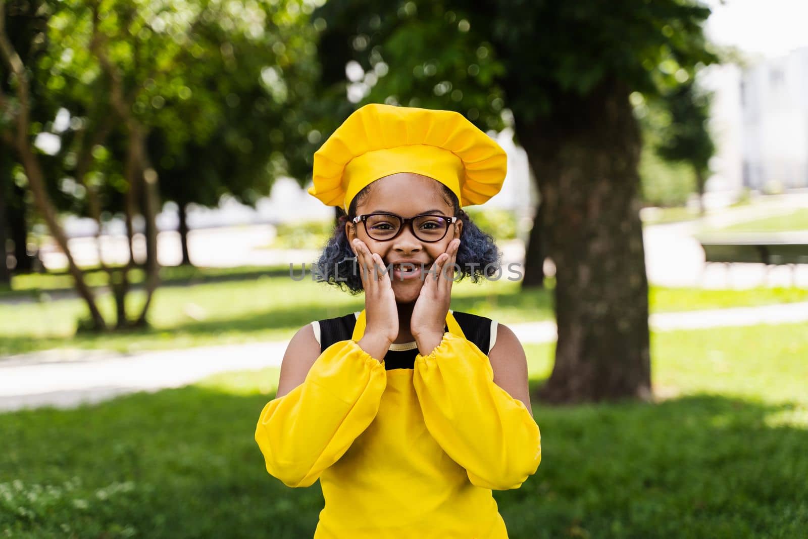 Shocked black african child cook girl in chefs hat and yellow apron uniform hold cheeks and surprise. Creative advertising for cafe or restaurant