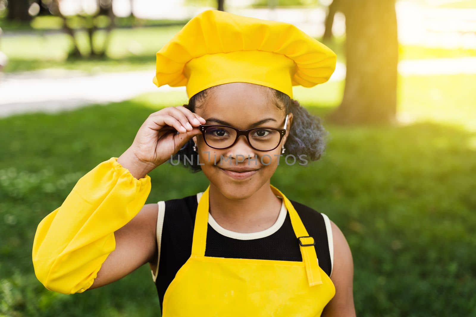 Black african child cook girl in chefs hat and yellow apron uniform touching hil glasses and smiling outdoor. Creative advertising for cafe or restaurant