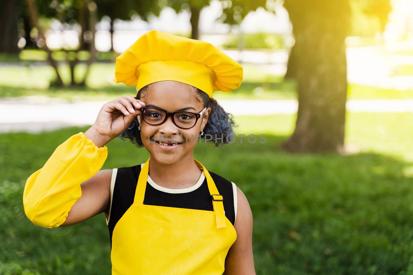 Black african child cook girl in chefs hat and yellow apron uniform touching hil glasses and smiling outdoor. Creative advertising for cafe or restaurant