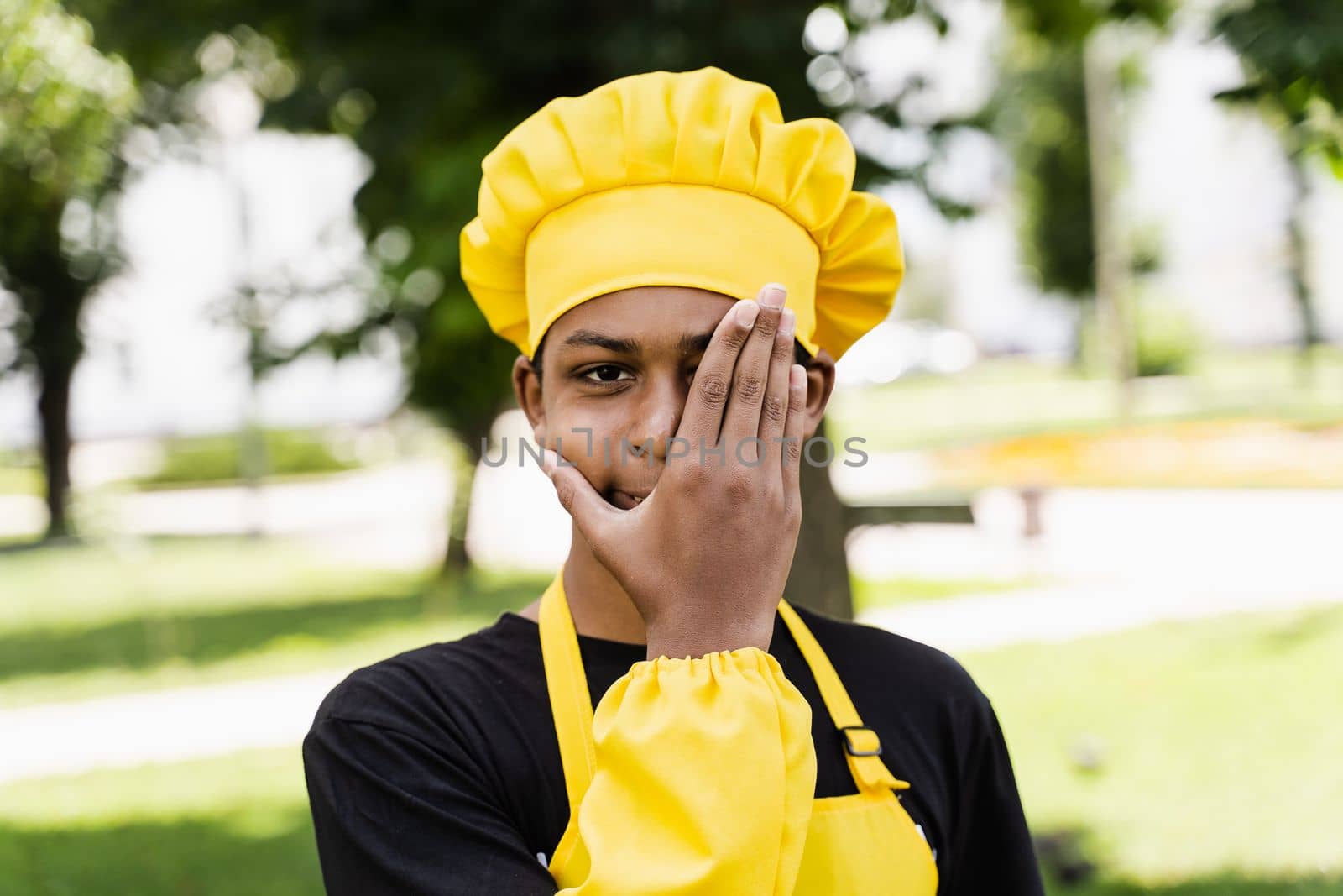 Black african teenager cook in chefs hat and yellow apron uniform hide face with hand. Creative advertising for cafe or restaurant