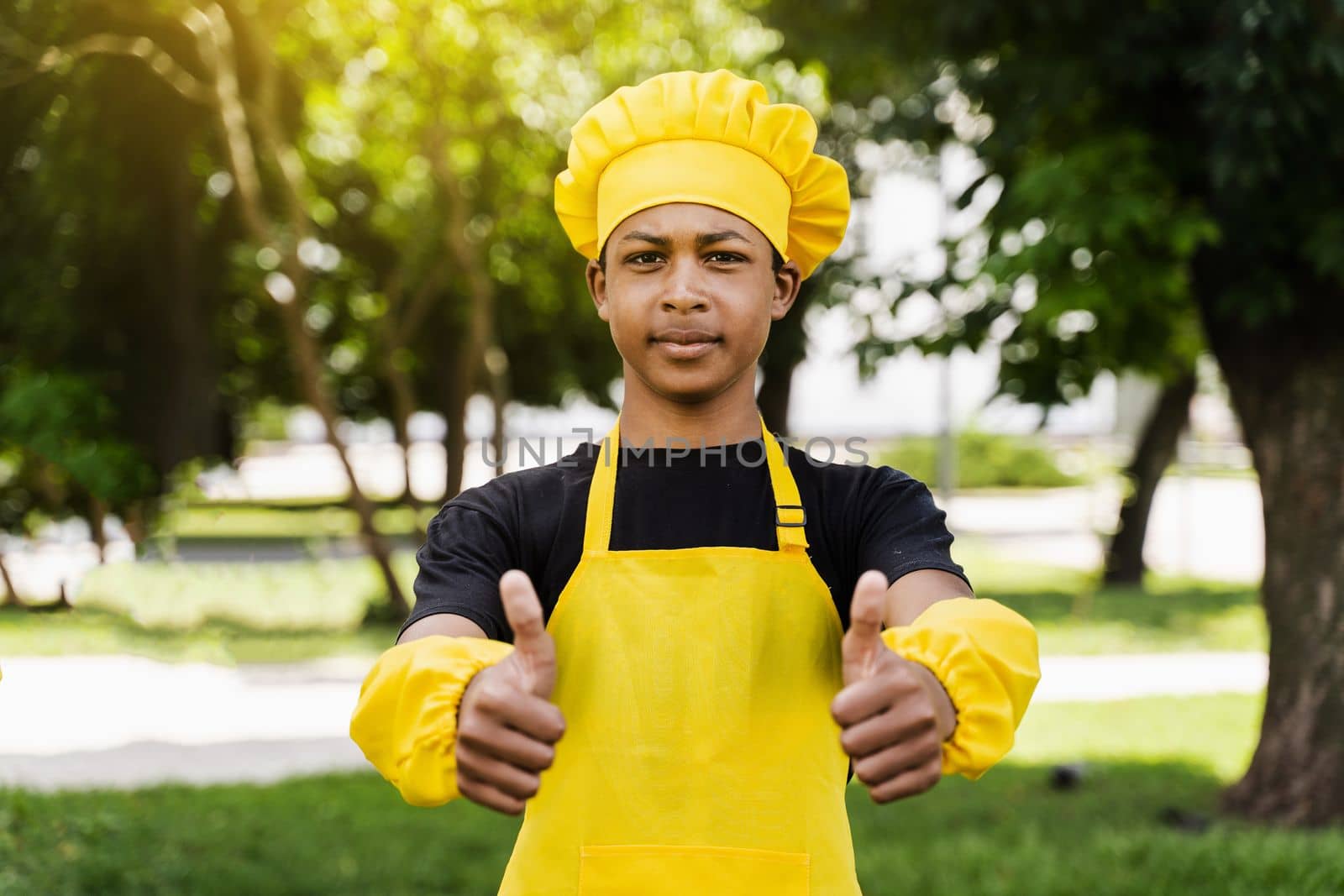 Black african teenager cook showing thumbs up in chefs hat and yellow apron uniform cooking dough for bakery. Creative advertising for cafe or restaurant