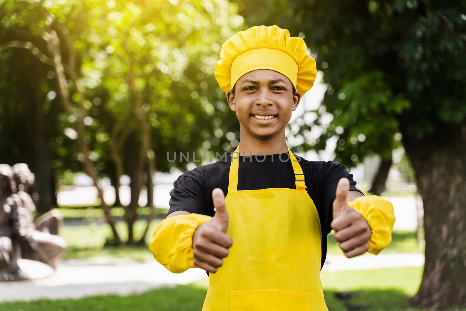 Black african teenager cook showing thumbs up in chefs hat and yellow apron uniform cooking dough for bakery. Creative advertising for cafe or restaurant
