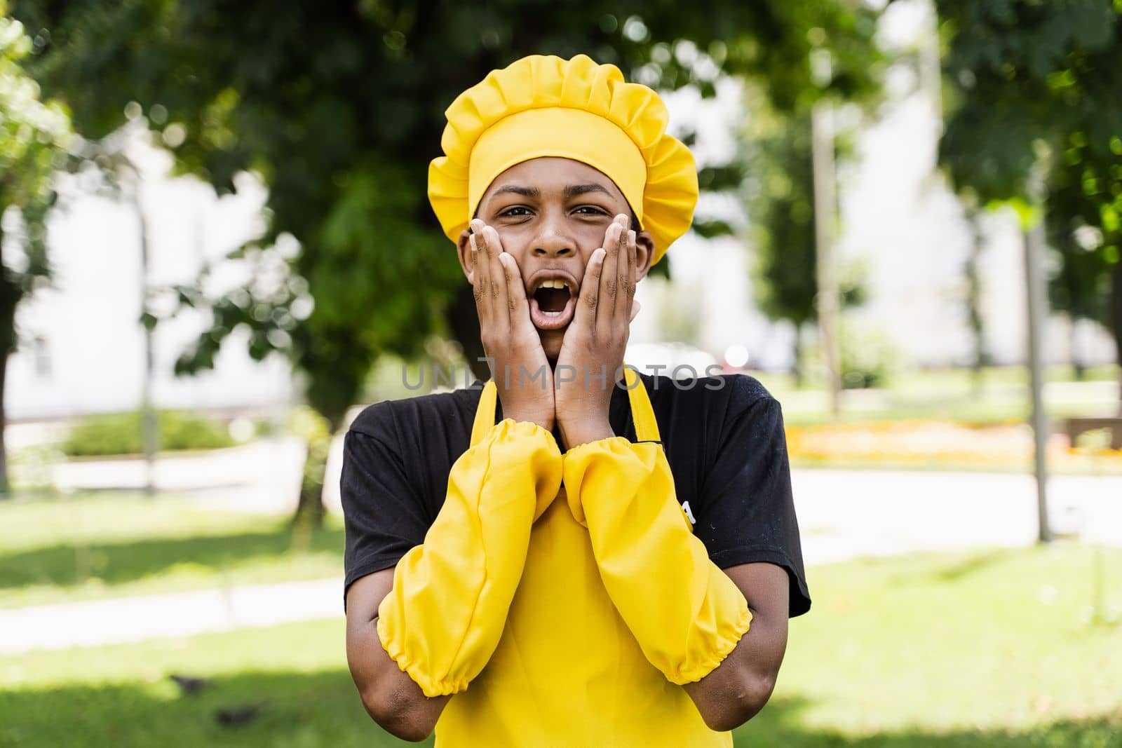 Shocked black african teenager cook in chefs hat and yellow apron uniform hold cheeks and surprise. Creative advertising for cafe or restaurant. by Rabizo