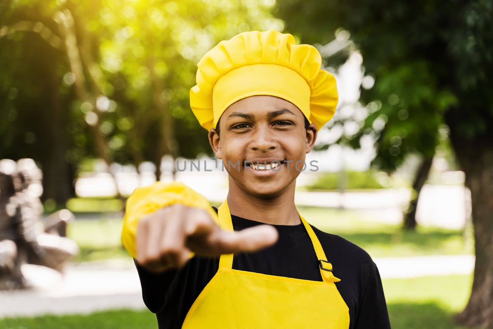 Handsome african teenager cook points to you. Black child cook in chefs hat and yellow apron uniform smiling and pointing to you outdoor. Creative advertising for cafe or restaurant.