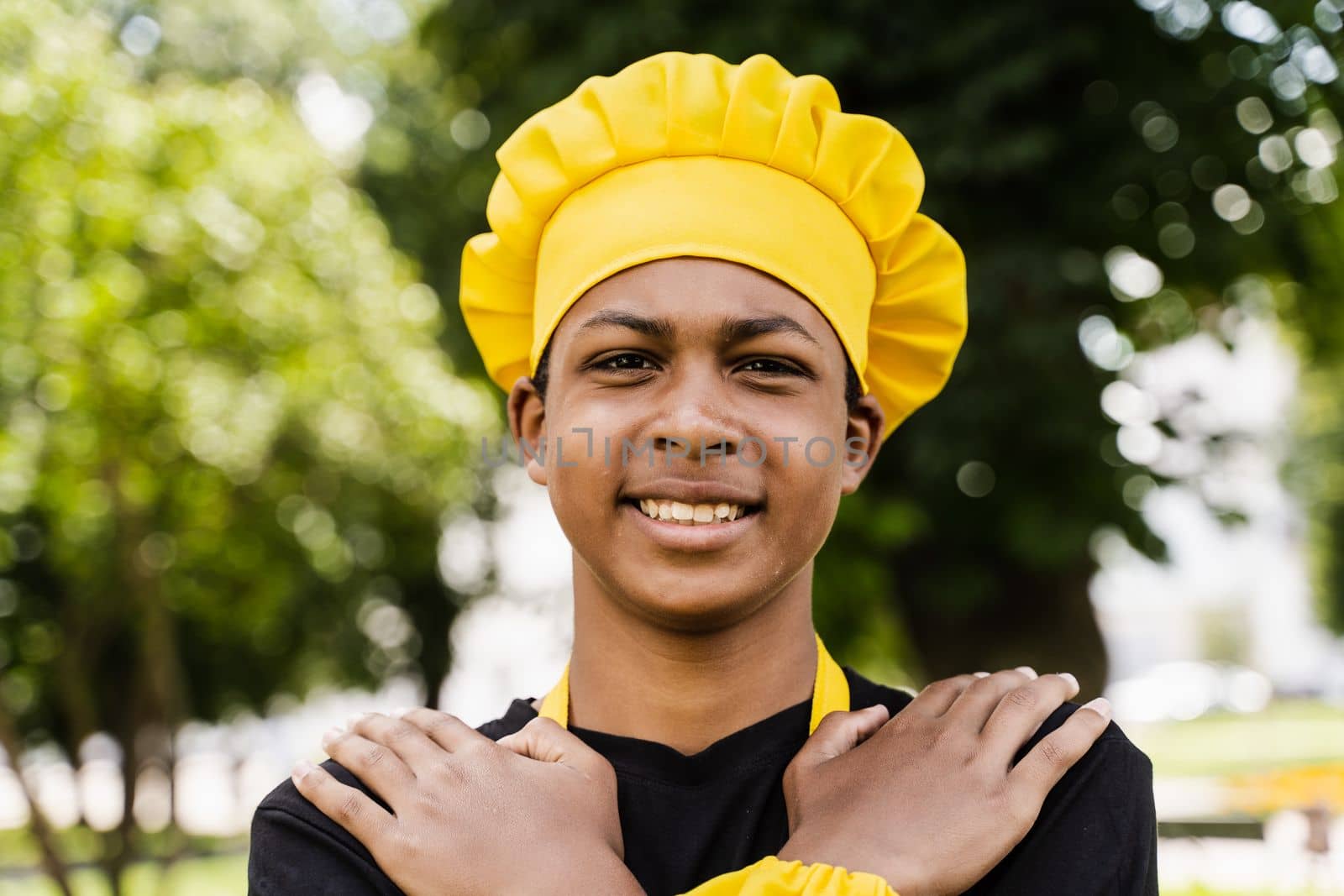 Black african teenager cook in chefs hat and yellow apron uniform smiling outdoor. Creative advertising for cafe or restaurant. by Rabizo