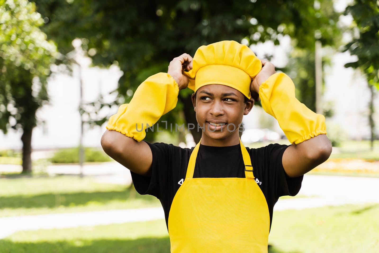 Black african teenager cook in chefs hat and yellow apron uniform touching his chefs hat. Creative advertising for cafe or restaurant
