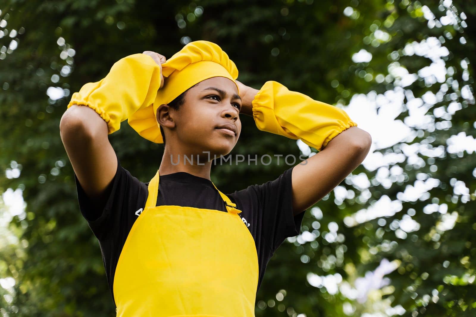 Black african teenager cook in chefs hat and yellow apron uniform touching his chefs hat. Creative advertising for cafe or restaurant. by Rabizo