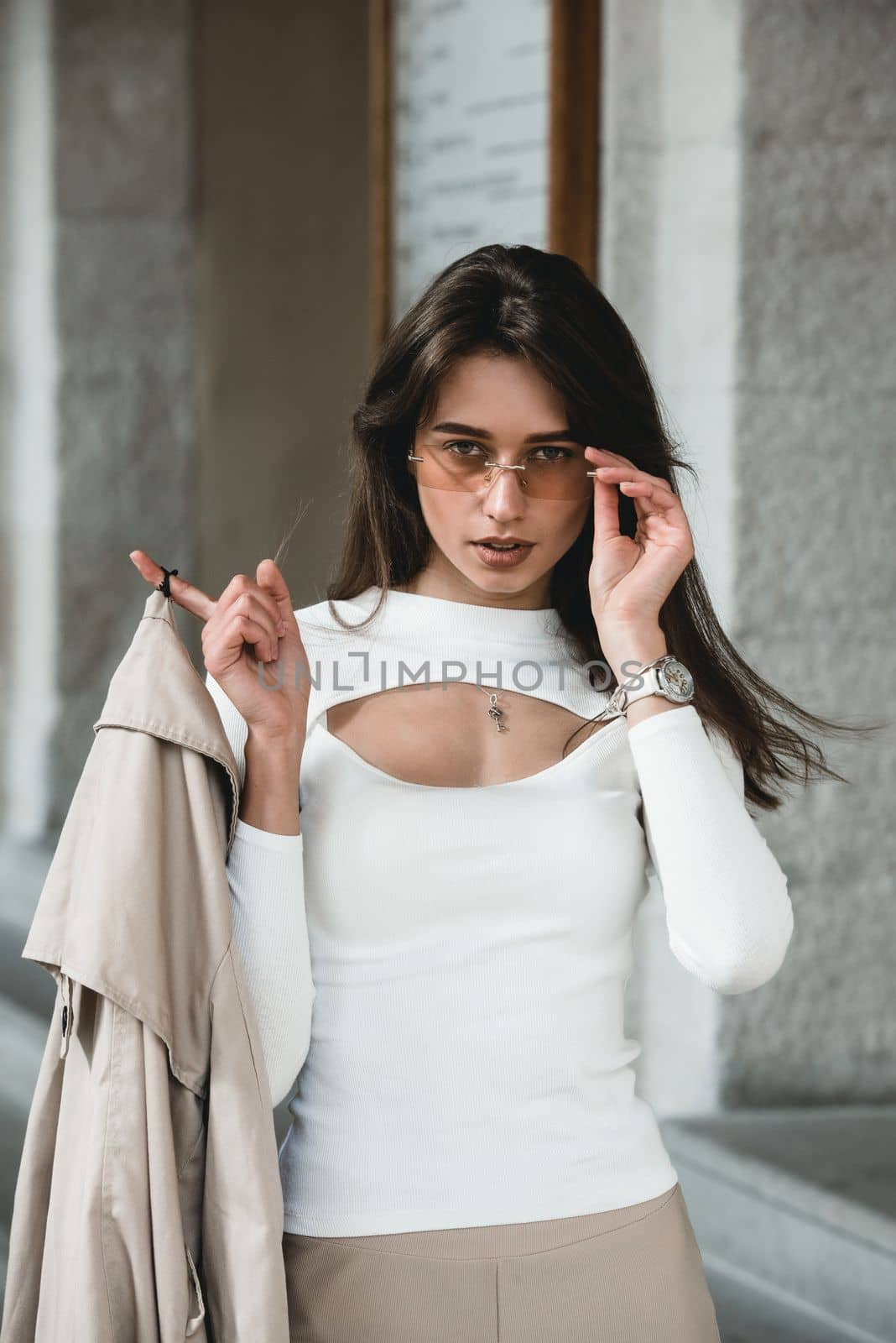 beautiful brunette girl in a sunglasses dressed in white blouse with a beige coat in hands. Stylish trendy fashion outlook.