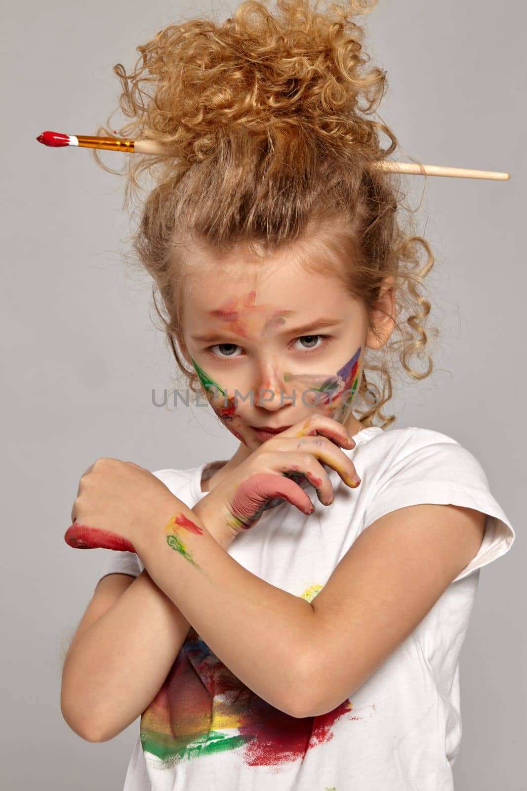 Beautiful little girl having a brush in her chic curly blond hair, wearing in a white t-shirt, has crossed her hands with a painted fingers, on a gray background.