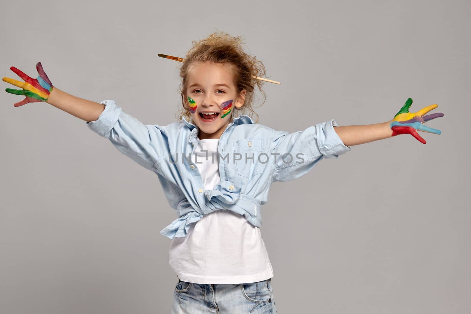 Beautiful little child having a brush in her chic hairstyle, wearing in a blue shirt and white t-shirt. She spread her arms widely and opened her mouth, looking at the camera on a gray background.