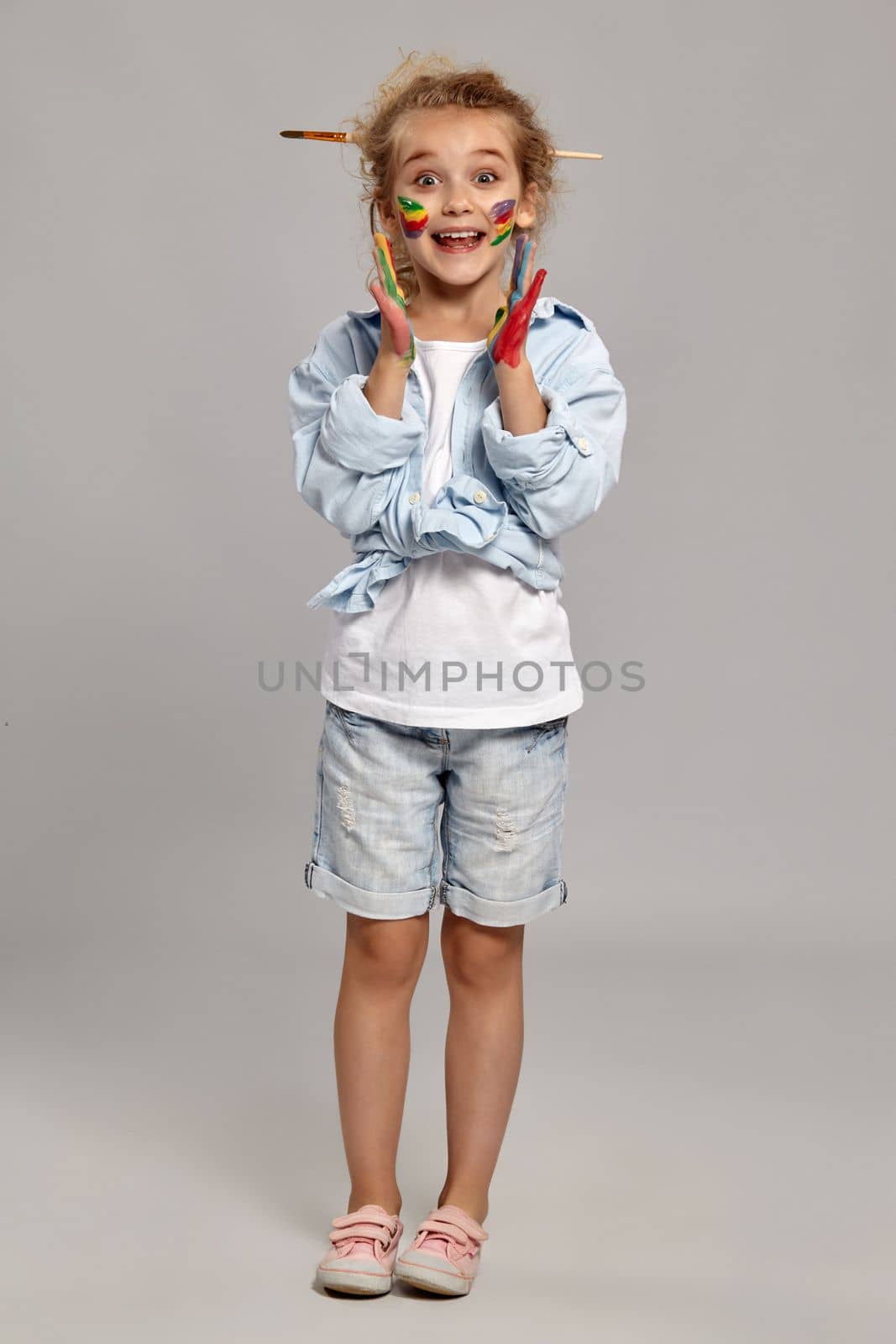 Cheerful child having a brush in her chic curly blond hair, wearing in a blue shirt and white t-shirt. She is posing with a painted hands and cheeks and looking wondered, on a gray background.