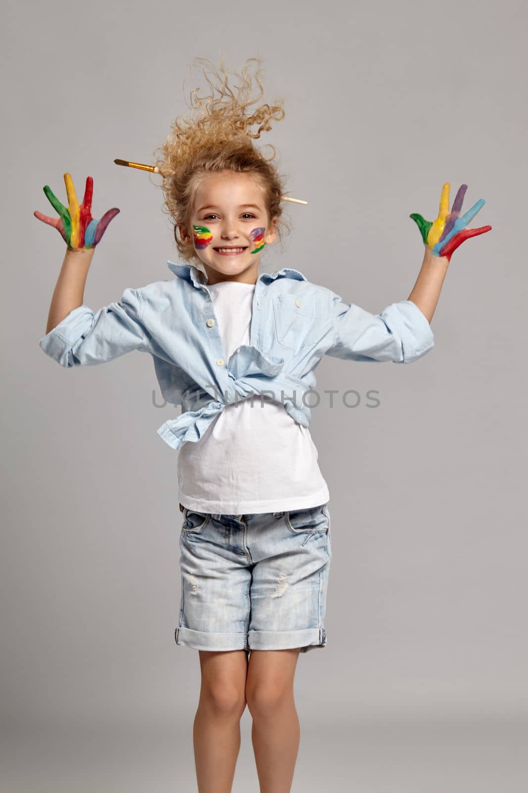 Cheerful girl having a brush in her chic curly blond hair, wearing in a blue shirt and white t-shirt. She raised her painted arms up and smiling, on a gray background.