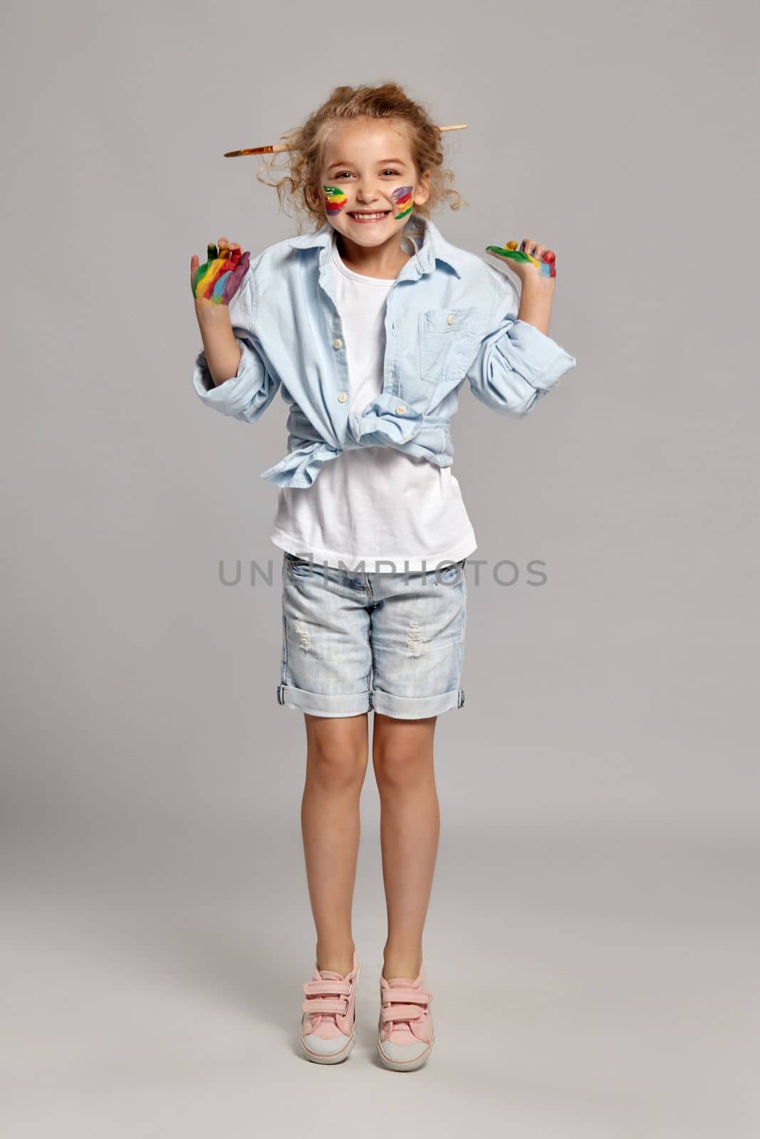 Cheerful little lady having a brush in her chic curly blond hair, wearing in a blue shirt and white t-shirt. She stood on tiptoe and smiling, on a gray background.