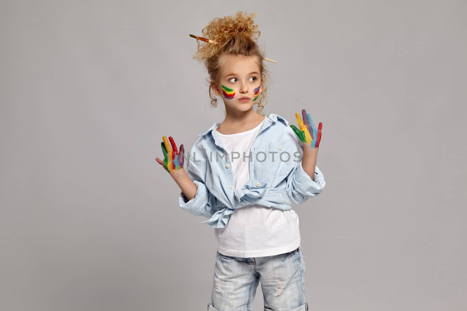 Lovely little girl having a brush in her chic hairstyle, wearing in a blue shirt and white t-shirt. She raised her painted hands up and looking away, on a gray background.