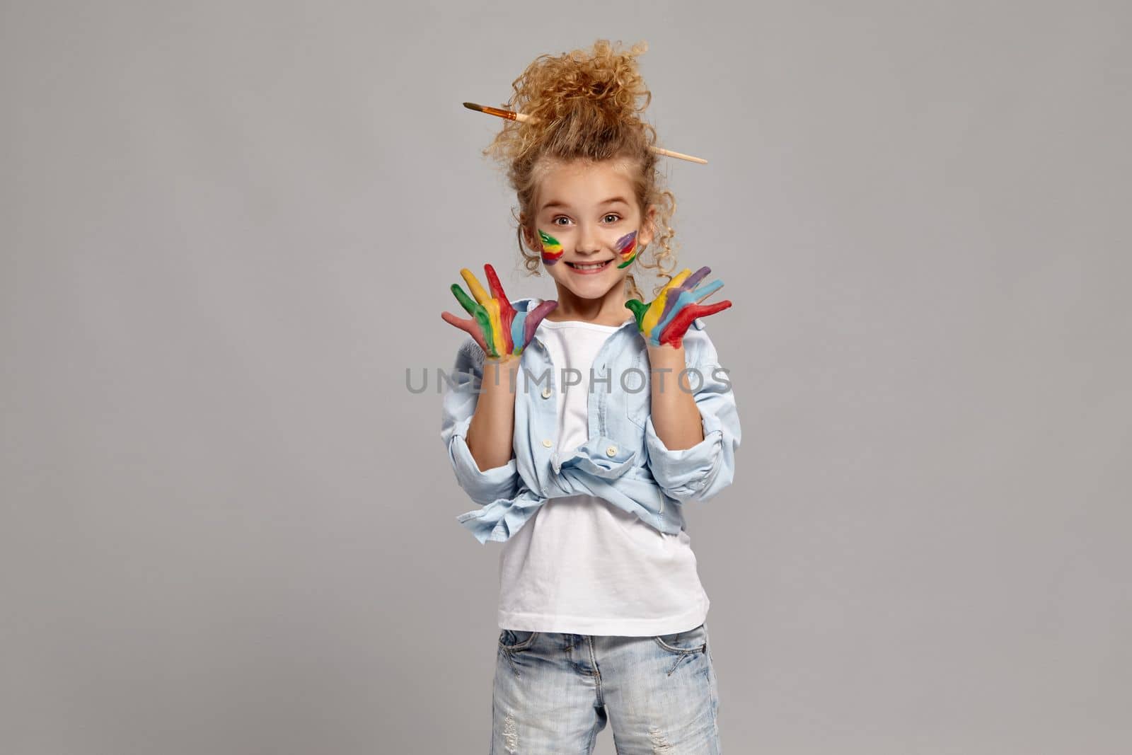 Lovely kid having a brush in her chic hairstyle, wearing in a blue shirt and white t-shirt. She raised her painted hands up, smiling and looking at the camera, on a gray background.